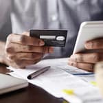 Close-up view of man's hands holding plastic credit card and smart phone. Dark-skinned businessman using mobile banking application on his gadget while paying bills at cafe online via internet